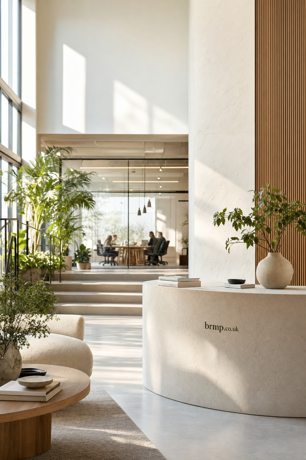 A bright, modern office reception with a curved stone desk featuring brmp.co.uk text, surrounded by plants and soft seating, leading into a glass-walled meeting room where four people are engaged in discussion.