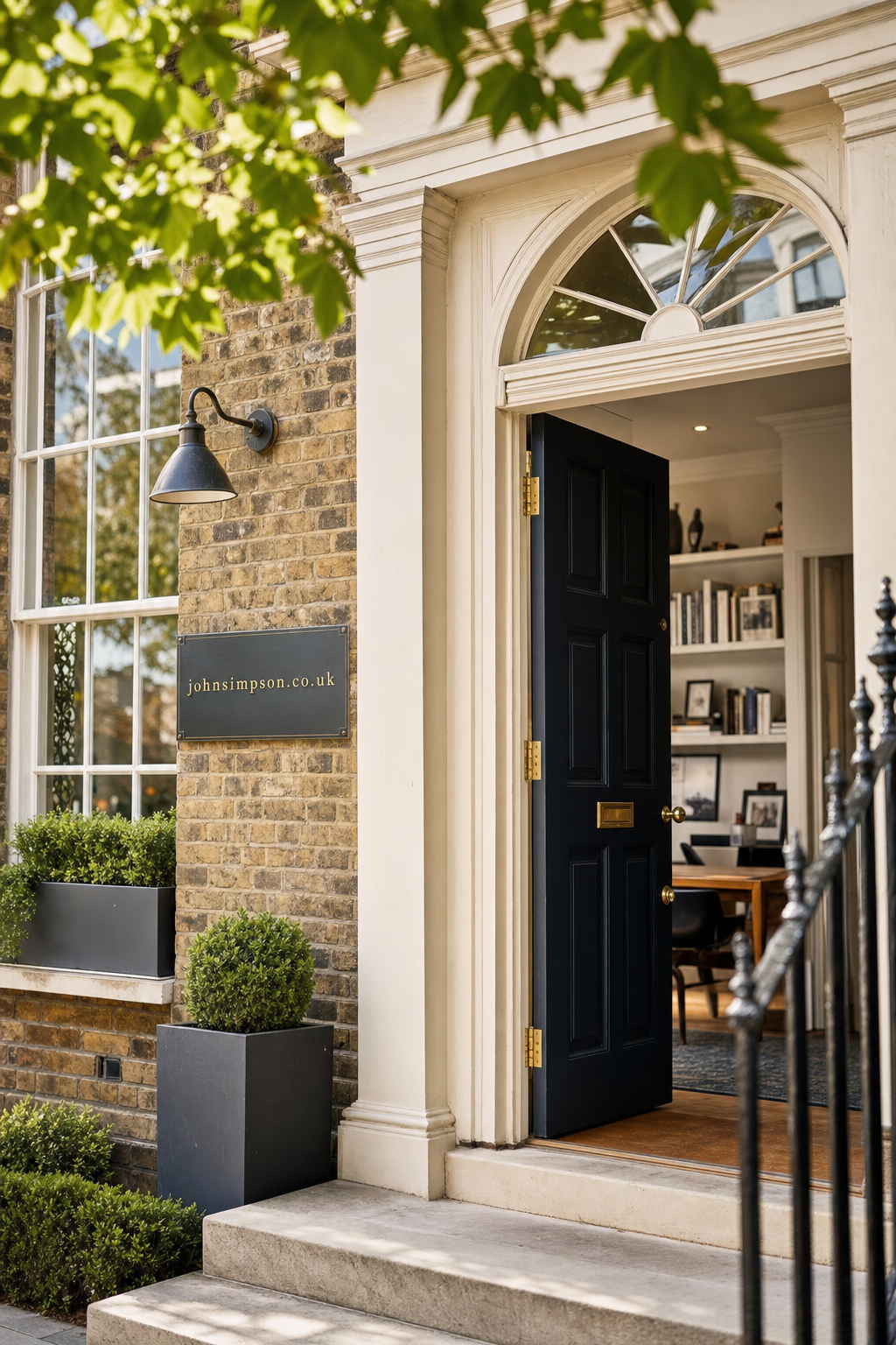 The welcoming entrance of a professional office with an open dark blue door, brick exterior, potted plants, and a brass sign displaying johnsimpson.co.uk beside the doorway under soft green tree leaves.