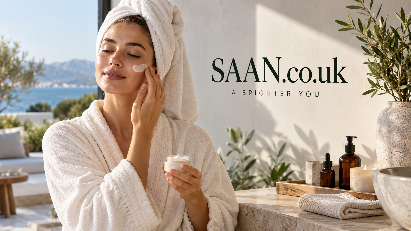 Woman in a white spa robe applying face cream beside natural skincare products, with warm sunlight, olive greenery and a coastal mountain view behind SAAN.co.uk branding.