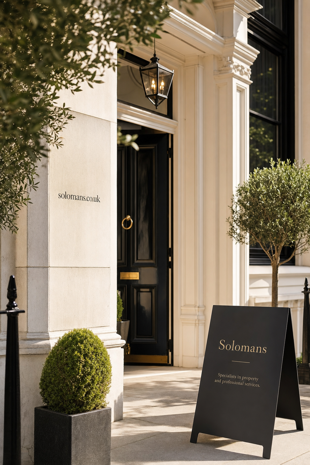 Entrance to a classic office building with a black door and gold handle, flanked by architectural columns and greenery; featuring a black sandwich board sign reading 'Solomans Specialists in property and professional services'
